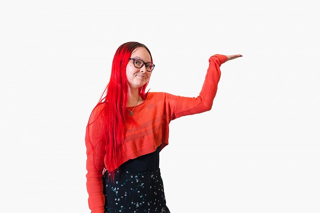 Henkilö katsoo kameraan ja pitää toista kättään ilmassa kuin esitelläkseen jotakin vierellään.